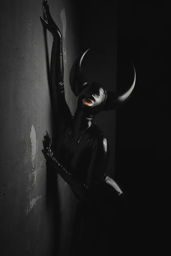 A figure in black latex with curved horns leans against a dark wall.