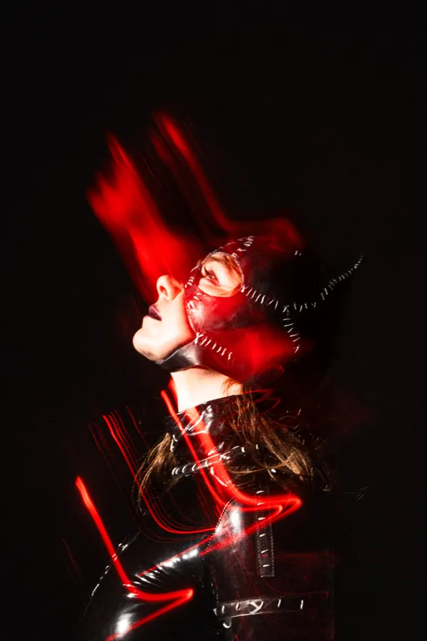 A woman in black latex with red horns, lit by a red light that blurs as she moves.