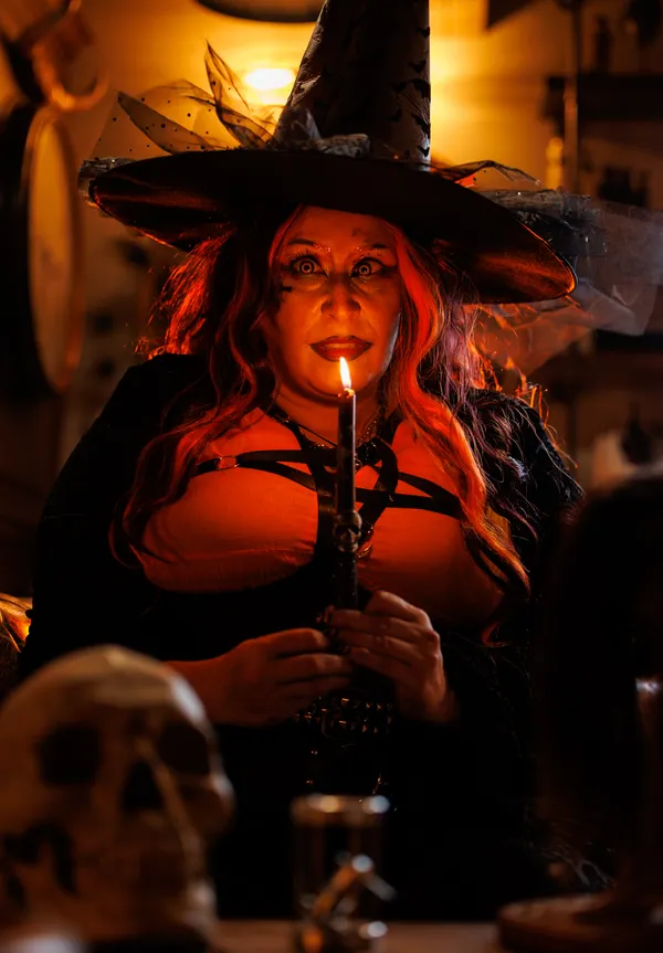 A witch in a black hat and leather harness holds a lit candle, a skull beside her.