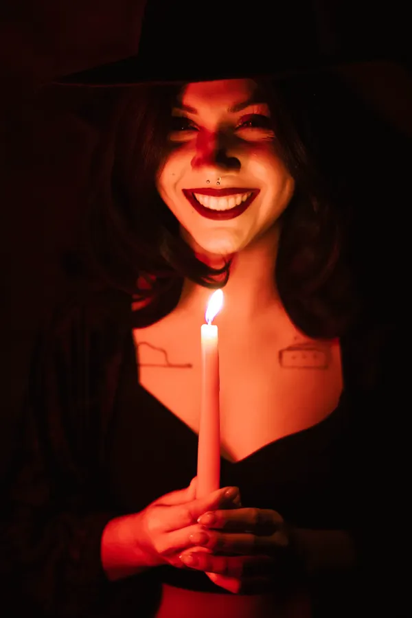 A smiling witch in a black hat holds a candle that lights her face red.