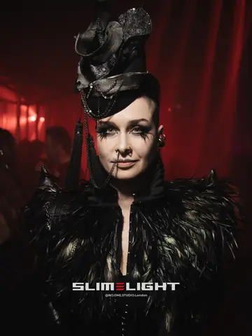 A guest in a black feathered jacket and a sculpted hat under red light.