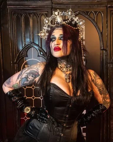 A tattooed guest in a black corset and a crown of crystals, in front of carved wooden panels.